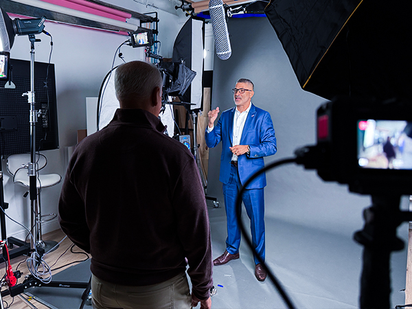 Man in a blue suit gives a talk on a video set as crew films him with cameras and lights nearby