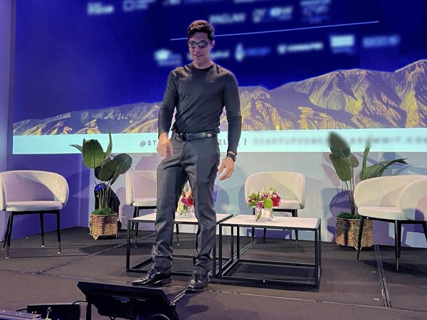 Man in a black long-sleeve shirt and jeans standing on a stage with small tables, chairs, plants, and flower arrangements against a blue backdrop with mountains.