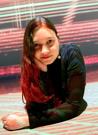 Portrait of a woman with long dark hair wearing a black top, resting her chin on her hand against a striped background.