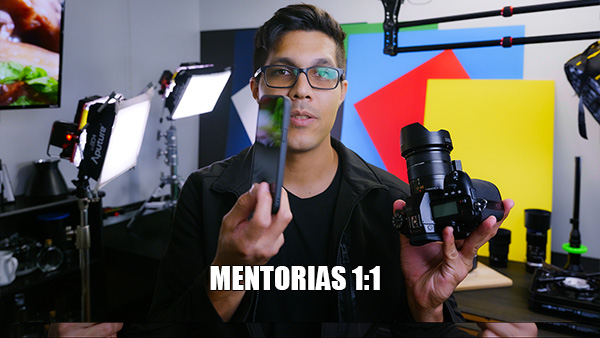 In a photography studio a man holds a smartphone in one hand and a camera in the other, with colorful backdrops and lighting gear visible.