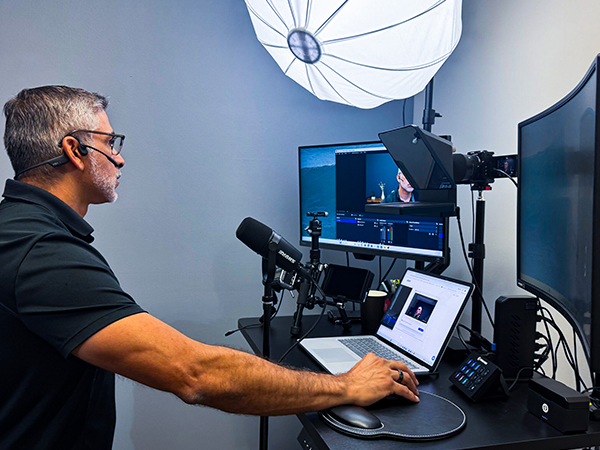 Man wearing a headset sits at a desk in a streaming studio with multiple monitors, a microphone, and a laptop.