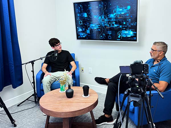 Two men in a podcast recording room: one speaks into a mic while the other watches, with a camera and laptop nearby and a digital display on the wall behind them.