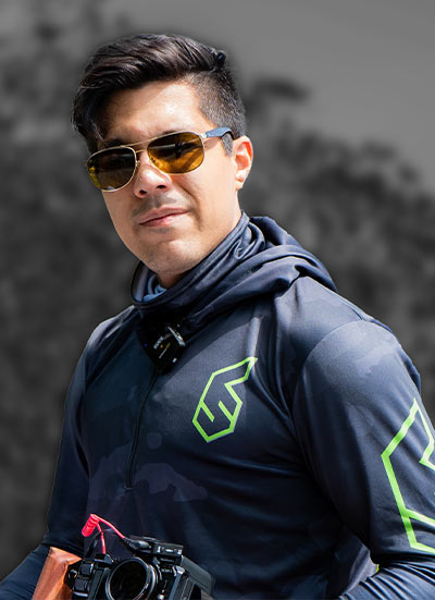 Photographer in a navy jacket with a neon logo, scarf, and orange sunglasses holding a camera outdoors.