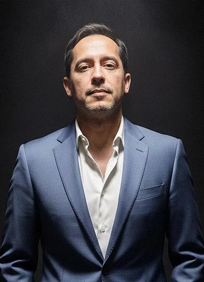 Man wearing a blue suit with a white shirt stands against a dark backdrop.