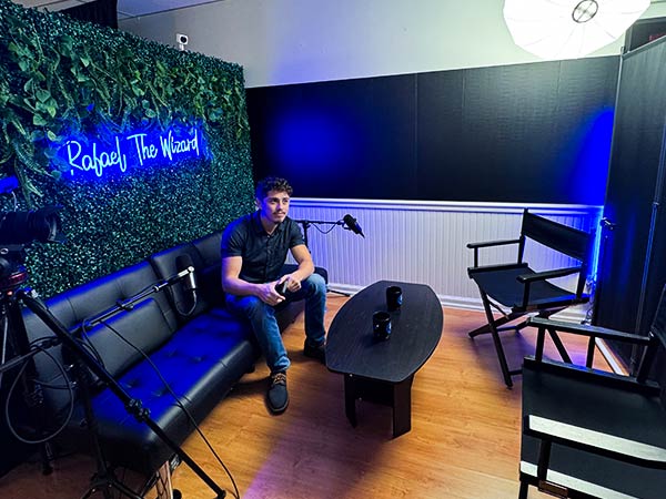 Man sits on a black sofa in a small recording studio with blue lighting, neon sign on a leafy wall, and camera gear nearby on a tripod and table with mugs nearby.