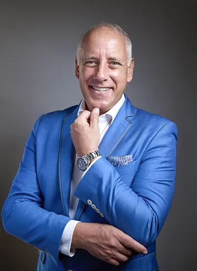 Smiling man in a blue blazer with a white shirt, posing with his hand on his chin in a professional headshot against a neutral background.